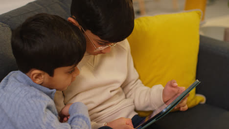 Two-Young-Boys-Sitting-On-Sofa-At-Home-Playing-Games-Or-Streaming-Onto-Digital-Tablet-10